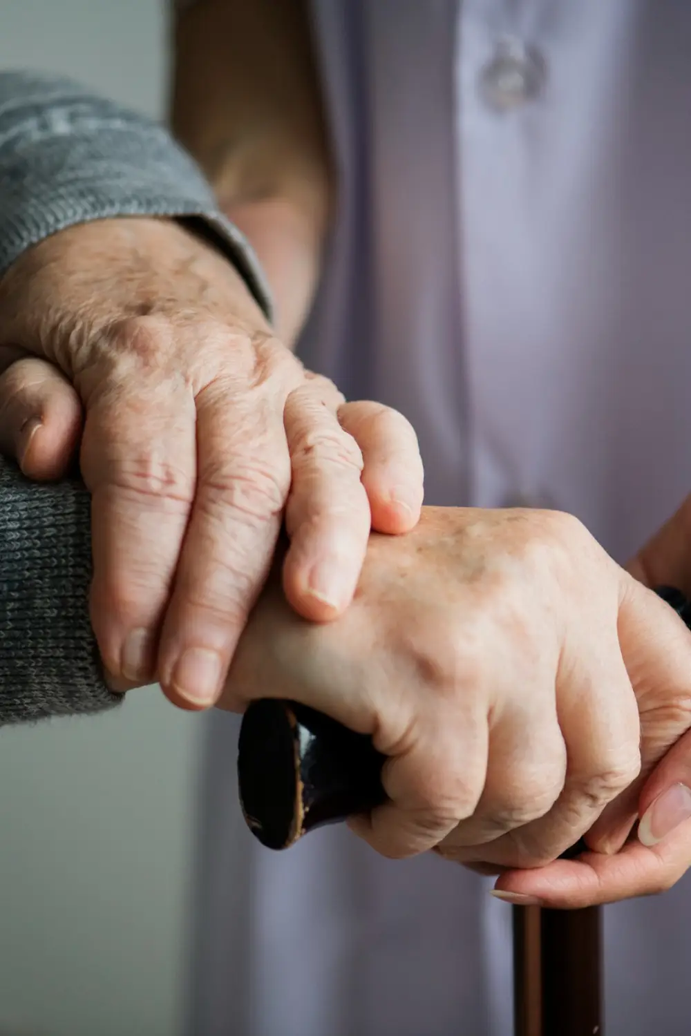 A caregiver gently holds the hand of an elderly person, who is gripping a cane, symbolizing support and assistance