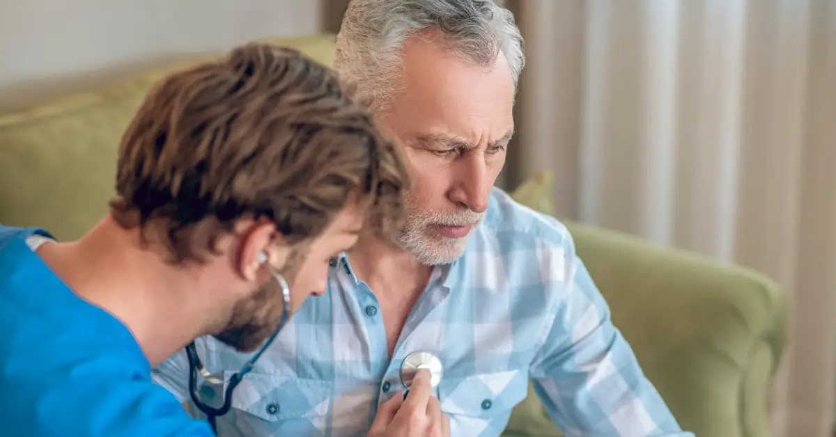A healthcare professional in blue scrubs uses a stethoscope to examine a patient sitting on a green couch in a cozy setting