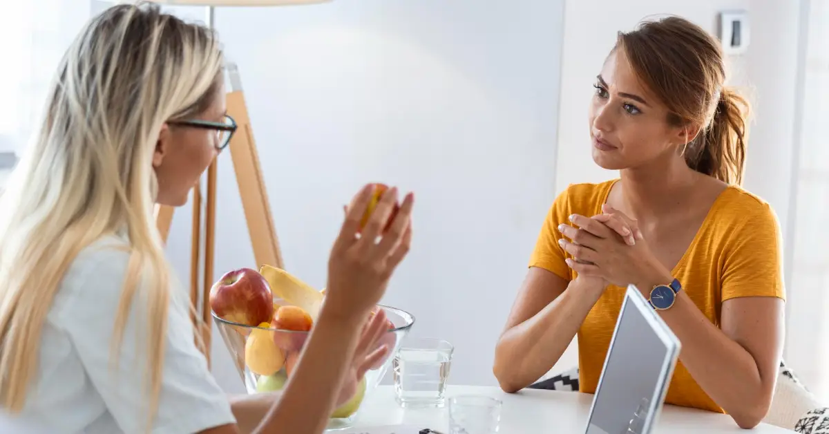Two women engage in a conversation at a table with fruits, a laptop, and a glass of water, in a bright, modern setting